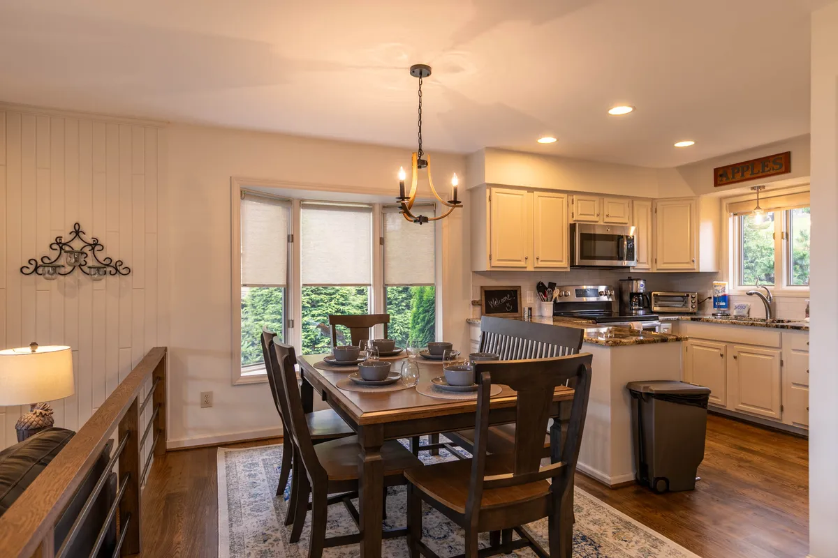 This image centers on a dining table beneath a chandelier, set near large windows that look out into the landscape, with the kitchen counter visible. The scene feels warm and welcoming, inviting guests to gather for meals while enjoying seasonal views through the windows.