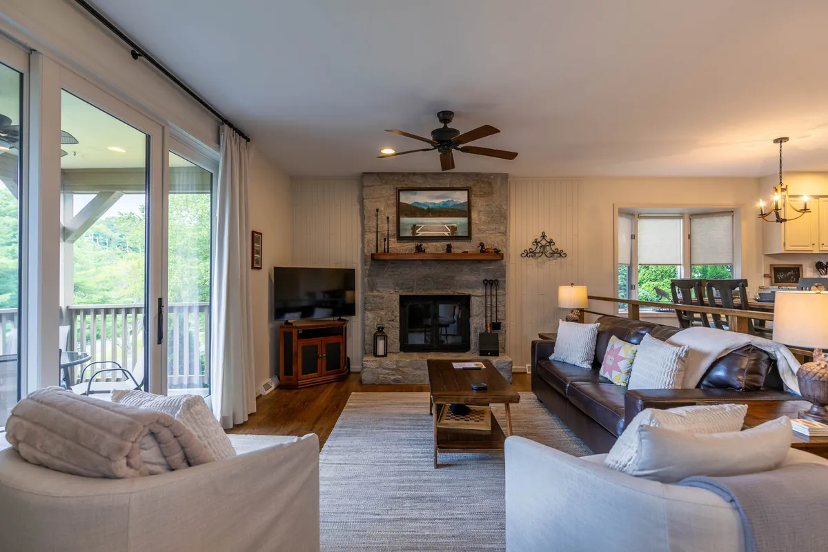 This image presents a panoramic view of the open living space. The scene showcases the fireplace as a cozy focal point, plush sofa seating, large windows that let in abundant natural light, and the adjoining dining area. The inviting arrangement encourages relaxation and social interaction, fitting the description of a welcoming, communal hub.