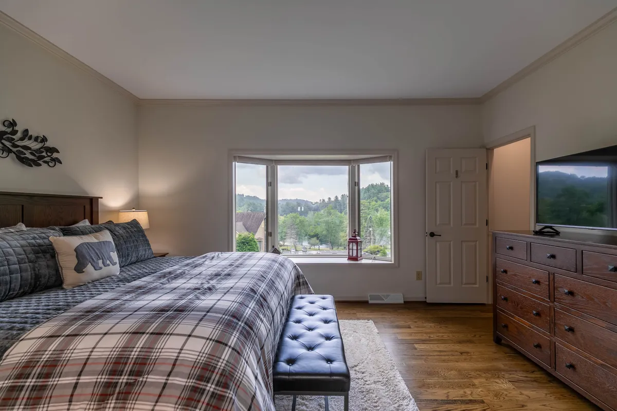 A bedroom featuring plaid bedding, a decorative bear pillow, a wooden dresser, and large windows framing a scenic view. The mood is cozy, ideal for relaxing or enjoying the scenery from bed.