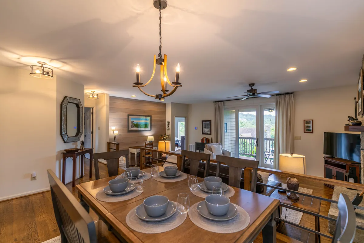 A well-lit dining space boasts a table set for a meal, a stylish chandelier, and a bright living area adorned with wall art. Natural light flows in, making the room feel open and connected to the outside. The setting encourages memorable dinners and peaceful mornings with coffee.