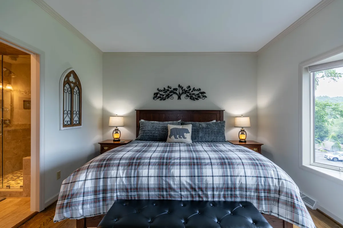 A comfortable king bedroom highlighted by plaid bedding, a whimsical bear pillow, nightstands with lamps, and a decorative wall piece. The space feels inviting and is designed for relaxation and quiet moments.