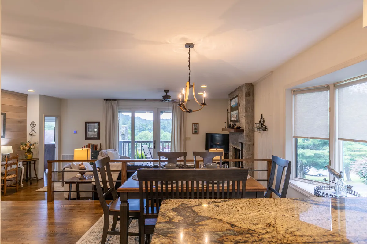 A dining and living area with a stone fireplace, large windows, and a balcony beyond. The space is lit warmly, perfect for cozy evenings, social gatherings, or relaxing while taking in the scenic environment outside.