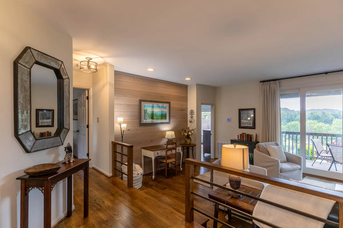 A cozy sitting area with a wooden accent wall, framed landscape painting, and comfortable chairs positioned near a table lamp. The scene is bathed in soft, natural light, creating an inviting atmosphere ideal for reading, working, or socializing. The balcony offers sunrise views that hint at a connection to the outdoors.
