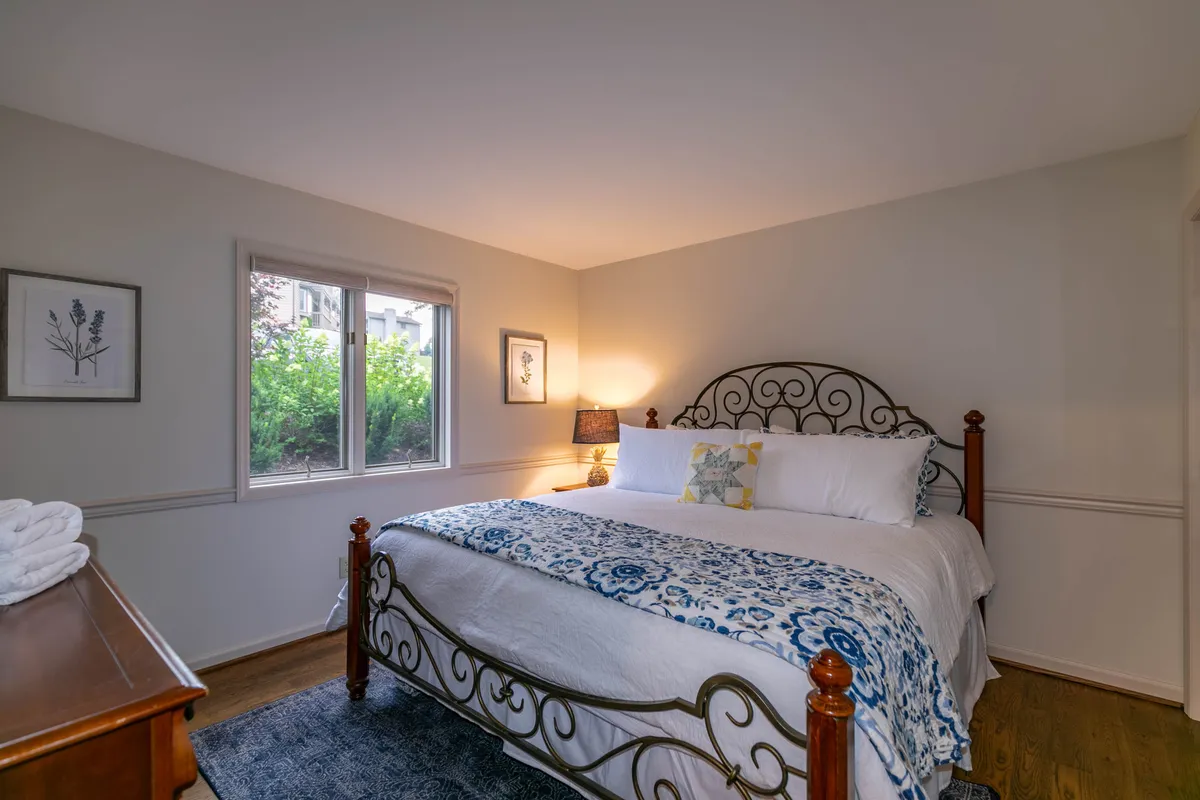 A serene king bedroom featuring a plush bed with decorative bedding, a nightstand with a lamp, and a window letting in ample natural light. The scene exudes a tranquil, cozy ambiance ideal for restful sleep or enjoying a quiet morning.